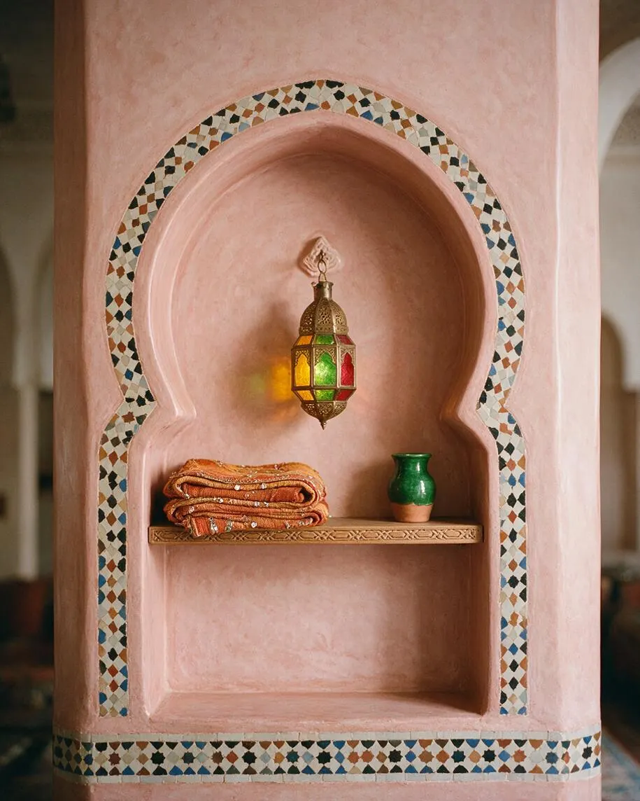 A Moroccan-style wall niche finished in soft pink plaster, framed by a multicolored mosaic arch and base border. Inside the alcove are a hanging brass lantern, folded patterned textiles on a carved shelf, and a small green ceramic vessel.