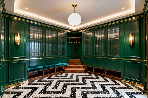 A glamorous Art Deco mudroom wrapped in emerald green paneled cabinetry with polished brass banding and pulls. Tall doors include reeded glass panels; a U-shaped walnut bench with open cubbies is topped by a teal velvet cushion. At the center, walnut steps rise to a corner fitted with a brass rail and hooks. The floor features a bold black-and-white terrazzo herringbone inset. Lighting combines fluted brass sconces, a white globe pendant, and soft ceiling cove LEDs.