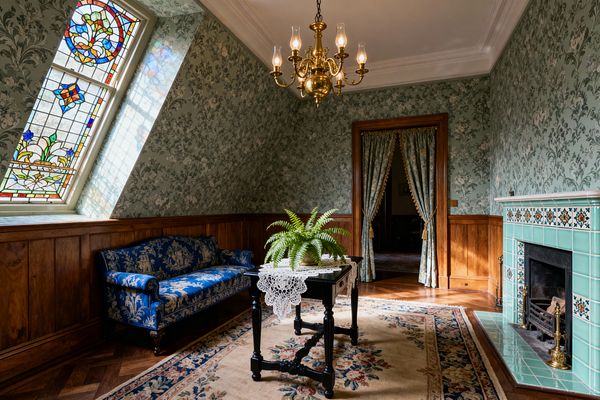A Victorian living room features sage floral wallpaper above oak wainscoting, a seafoam tiled fireplace with patterned inserts and brass tools, and a blue jacquard settee along a stained-glass dormer window. A black turned-leg table with a crochet lace runner and a fern sits on a floral wool rug beneath a brass chandelier, while tasseled damask curtains frame a wood-trimmed doorway.
