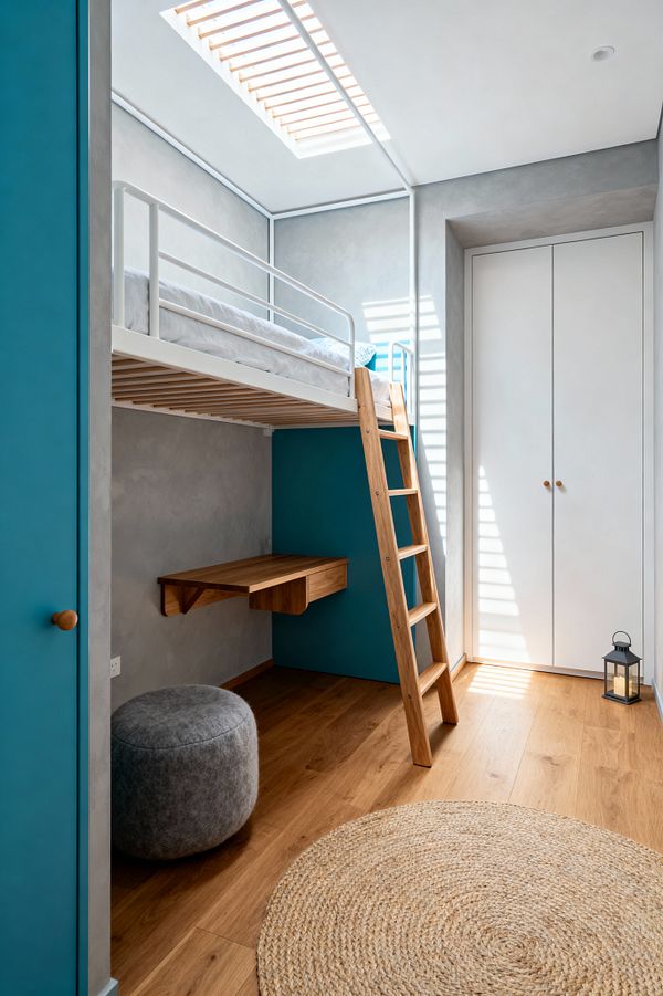 A minimalist kids’ room with a white loft bed and guardrail accessed by a natural oak ladder. Beneath the bed is a floating oak desk against a teal accent panel, with smooth gray plaster walls, oak flooring, a round jute rug, and a gray felt pouf; daylight streams from a skylight with timber slats, casting stripes across a white built-in wardrobe with wooden knobs and a small black lantern near the door.