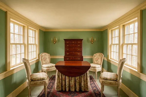 A traditional sunroom with sage green walls, cream trim, and large multi-pane windows on both sides that fill the room with natural light. Four upholstered floral chairs surround a polished mahogany game table with a skirted base, while a matching mahogany chest and brass wall sconces add formal elegance.