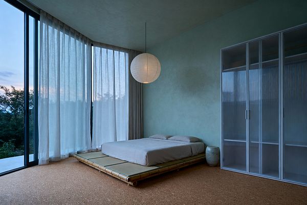 A minimalist bedroom shows a low tatami platform bed with a bamboo frame and gray linen bedding next to a small ceramic stool. Floor-to-ceiling corner windows with dark frames are draped in sheer white curtains, casting soft light on cork floors and pale seafoam limewashed walls. A round white paper lantern hangs centrally, and a slim wardrobe with fluted polycarbonate sliding doors lines the right wall, creating a calm, uncluttered space.