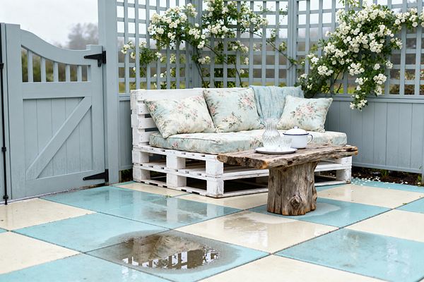 An outdoor patio enclosed by a duck-egg blue fence with lattice trellis features a DIY sofa made from white-painted pallets, layered with pastel floral cushions and a pale blue throw. A rustic live-edge stump table holds a glass carafe, a white enamel teapot with blue rim, and a tumbler. The floor is a checkerboard of large aqua and cream pavers with rain puddles. Climbing white roses trail along the trellis, with a matching gate to the left.
