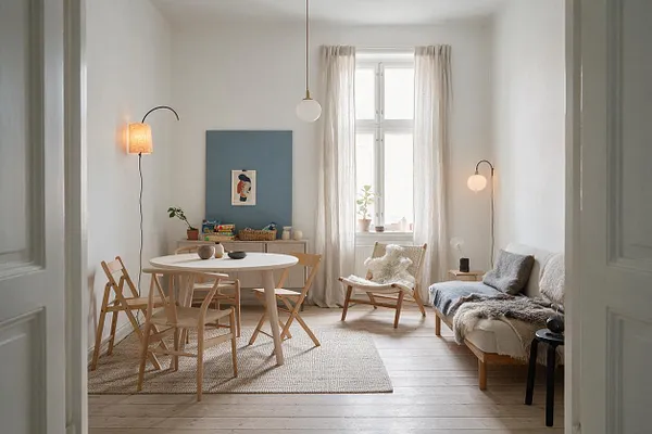A bright Scandinavian-style game room with white walls, pale wood floors, and soft natural light from a tall window with sheer curtains. The room includes a round white table, light wood chairs, a cozy bench with gray and white throws, a woven rug, wall sconces, and a blue accent panel with small decor and games.